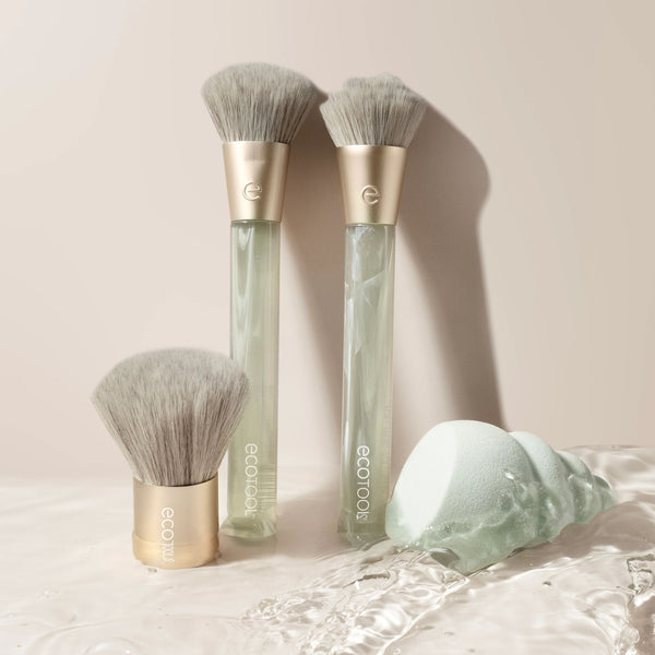 Face brush set on light surface: three brushes with gold tops, clear handles, sponge