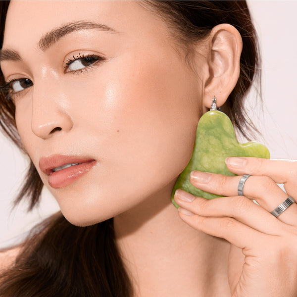Green jade gua sha held to the cheek for facial massage, heart-shaped stone