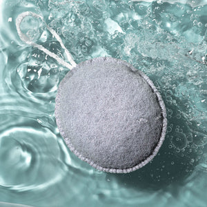 Grey oval exfoliating puff with stitched edge floating in water bubbles