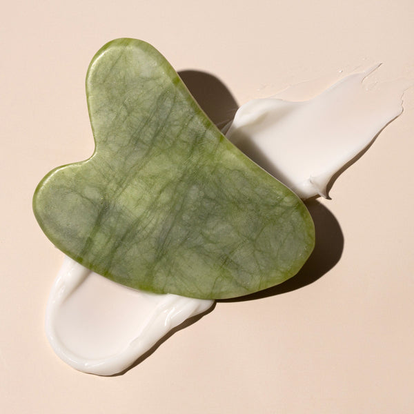 Heart-shaped green jade gua sha stone lying on white cream